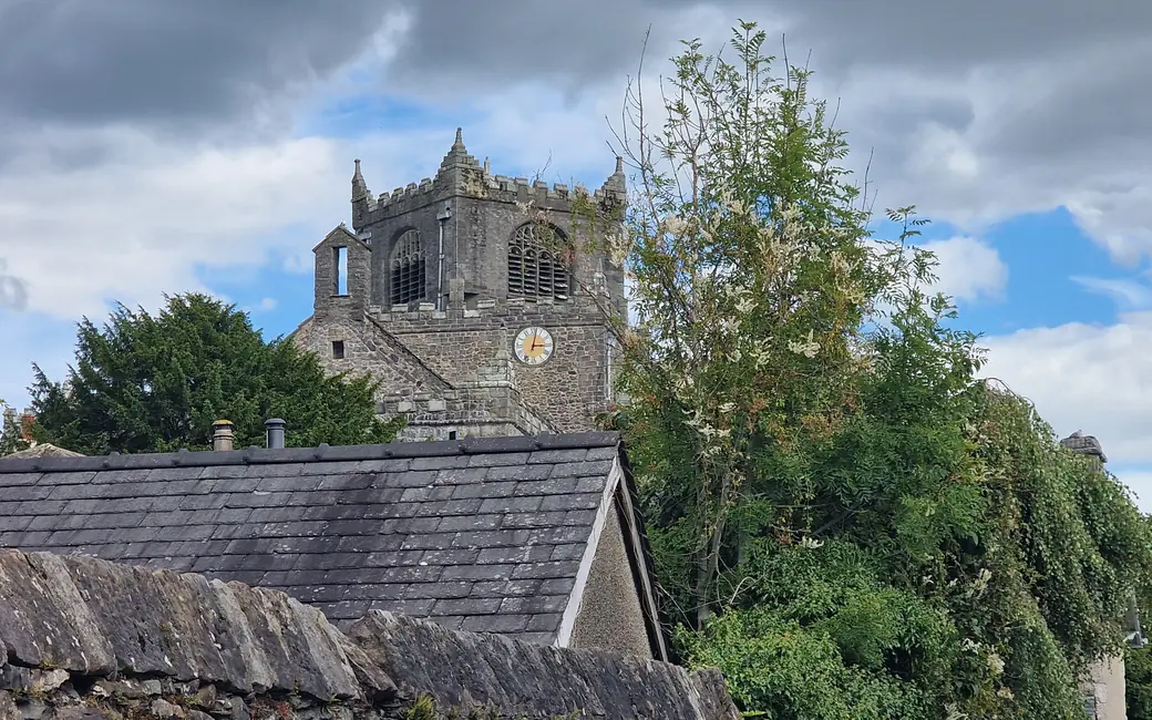 The battlemented tower of Cartmel Priory with its clock face rising above slate rooftops and a drystone wall